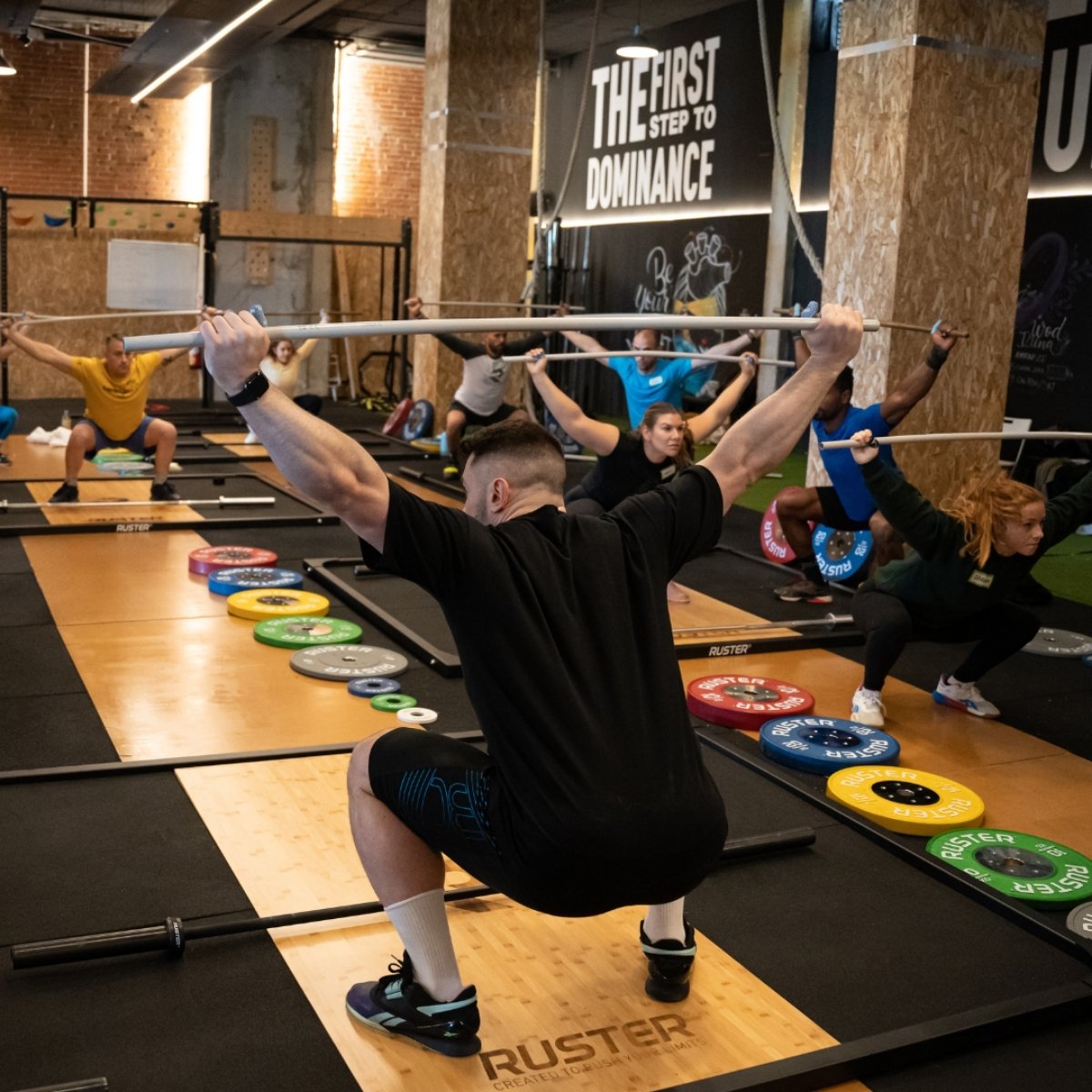 A group of people, in a gym setting, perform synchronized overhead squats with barbells (Torokhtiy Weightlifting)