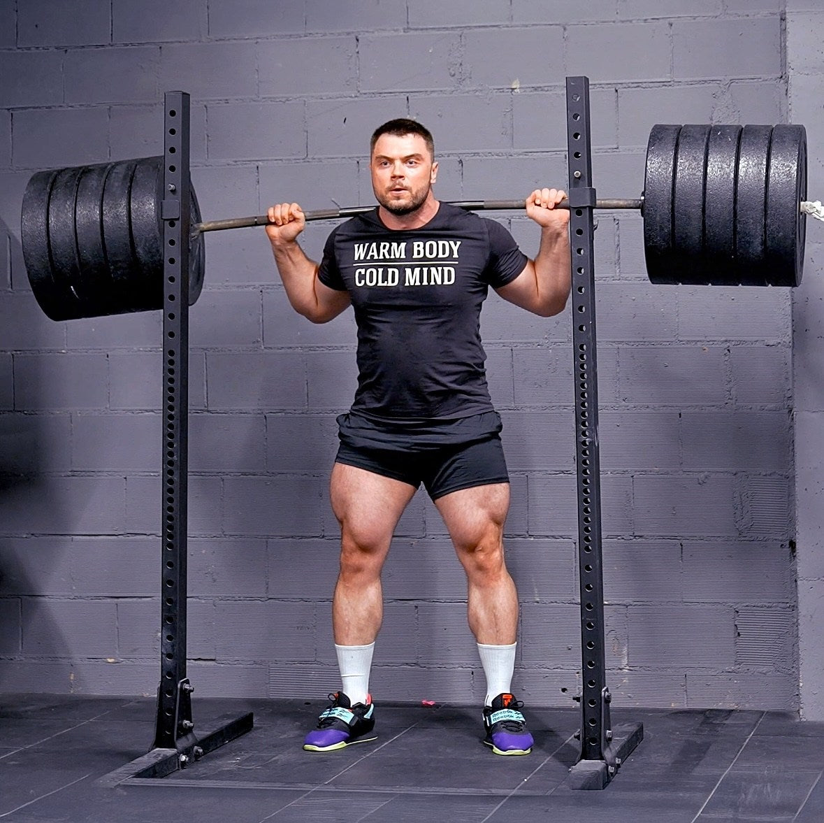Man performing a barbell squat in a gym setting with a gray brick wall background. (Torokhtiy Weightlifting)