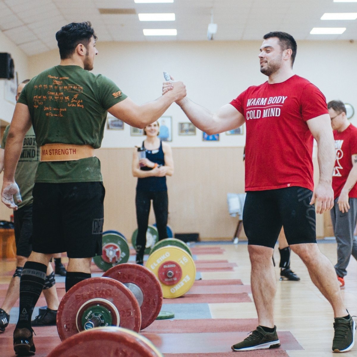 Two men shaking hands in a gym setting with weightlifting equipment in the foreground (Torokhtiy Weightlifting)
